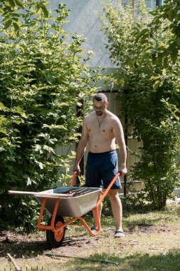 Mavi şort giyen üstsüz bir adam yaz boyunca sık yeşil çalıların arasında bir el arabasını iterek sıradan bir bahçe hayatı sahnesi yakalıyor.