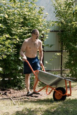 Mavi şort giyen üstsüz bir adam yemyeşil çalıları ve ahşap çitleri olan bir bahçe arazisinde turuncu bir el arabasını itiyor. bu resim açık hava çalışmalarını betimler