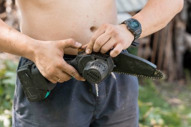 Tişörtsüz bir adam açık havada yemyeşil bitki örtüsünün ortasında dikilirken bir bataryayı kablosuz testereye titizlikle bağlıyor.