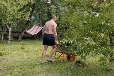 Tişörtsüz bir adam, portakal renkli bir el arabasını meyve ağaçları ve yeşilliklerle dolu çimenli bir bahçeye itiyor. Pembe çizgili bir hamağın yanında, bir yaz bahçesi çalışma sahnesini temsil ediyor.