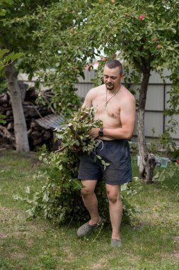 Şortlu üstsüz bir adam canlı bir bahçede elma ağacıyla ilgilenir. Bir odun yığınının yanında budama dalları, kırsal yaşamı resmediyor, yaz bakımını yapıyor ve rahat bir ruh hali içinde.