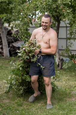 Tişörtsüz bir adam dışarıda bahçeyle uğraşıyor, arka bahçesinde bir elma ağacı ve odun yığınıyla dallar taşıyor. Sıradan yaz yaşam tarzı, gevşemiş ruh hali, yeşil çimenler.