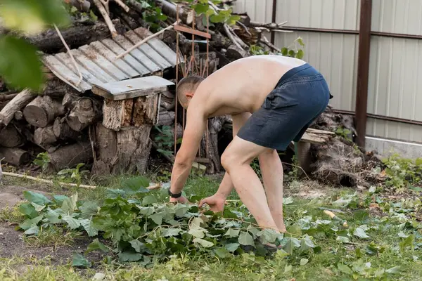 Tişörtsüz bir adam, bahçeye eğilmiş, sarmaşıklarla dolu, yıpranmış bir odun yığını ve güneşli arka bahçedeki galvanize metal çitin yanında.