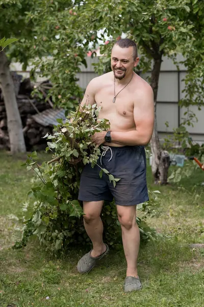 Tişörtsüz bir adam dışarıda bahçeyle uğraşıyor, arka bahçesinde bir elma ağacı ve odun yığınıyla dallar taşıyor. Sıradan yaz yaşam tarzı, gevşemiş ruh hali, yeşil çimenler.