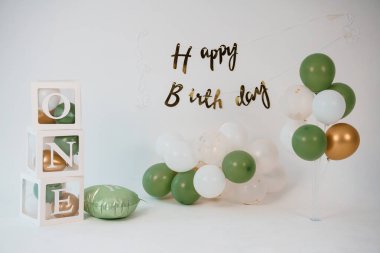 Ahşap bir blok, yeşil ve beyaz balon çelengi ve konfetiden oluşan sevimli bir ilk doğum günü düzeneği, bebek için bir dönüm noktası oluşturan neşeli ve kutlayıcı bir atmosfer yaratıyor.