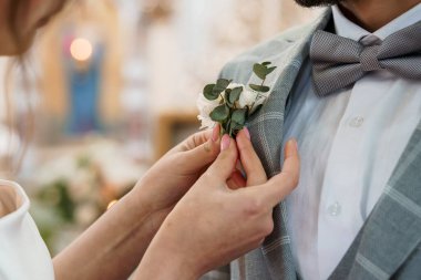 Yakın plan bir gelinin okaliptüs ile narin bir yaka çiçeğini damatların yakasına yerleştirdiği bir düğün töreninde gri bir takım elbise giydiğini gösteriyor.