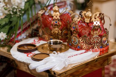 Süslü altın bir Ortodoks düğün tacı, ayin mumları ve kırmızı tekstil ve beyaz çiçeklere karşı dekore edilmiş altın bir masanın üzerinde küçük bir tekne.