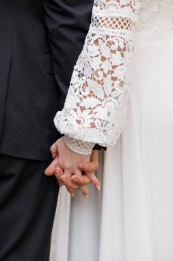 Bir düğün çifti el ele tutuşurken, siyah bir takım elbise ve karmaşık beyaz dantelli gelinlik sergilerken, aşkı, bağlılığı ve samimiyeti simgeleyen yakın çekim.