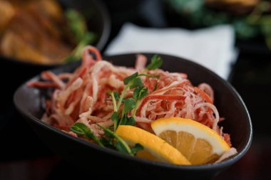 Yakın plan çekimde canlı bir Japon yengeç salatası resmedilir. Limon ve mikro yeşilliklerle süslenmiş koyu bir çömlek kabında güzelce sunulur.