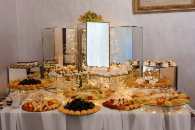 Ayna sütunları, meyveler ve pastalarla dolu katmanlı tabaklar, yumuşak beyaz çarşaflar ve düğün ve kutlamalar için sıcak dekoratif ışıklandırma içeren lüks bir tatlı masası.