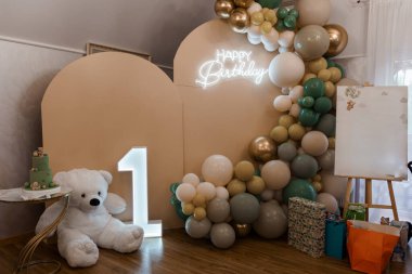 Çekici bir ilk doğum günü düzeninde 'Mutlu Yıllar' işareti, yeşil ve altın tonlarında bir balon çelengi, büyük bir '1' numarası ve sevimli bir oyuncak ayı bulunur. Parti davetleri ve sosyal medya grafikleri için ideal