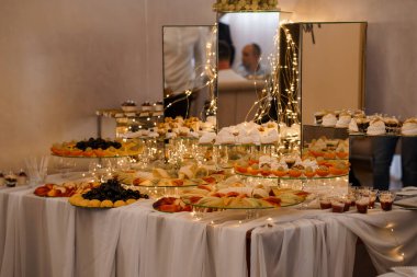 Müsrif bir düğün tatlı büfesi çeşitli meyve, kek ve hamur işlerini sicim ışıkları ve aynalarla süslenmiş tabaklarda sergiliyor.