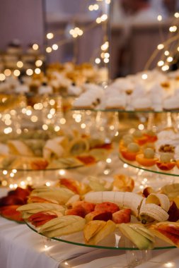 Göz alıcı peri ışıklarıyla süslenmiş cam katlı bir masada güzel bir şekilde hazırlanmış meyve tabağı ve lezzetli pastalar düğün resepsiyonları, partiler ve şenlikli kutlamalar için mükemmel.