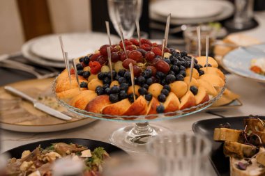 Üzüm, böğürtlen, şeftali ve kavun sergilenen güzel bir meyve tabağı resmi bir masadaki aperatiflerin yanında oturmuş kutlama yemeği öneriyor.
