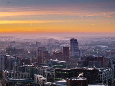Şafak vakti Leeds şehir merkezinin çarpıcı hava manzarası, şehir mimarisi, canlı renkler ve sisli arka plan tepeleri, modern büyümeyi, dingin ortamı ve mimari güzelliği vurguluyor..