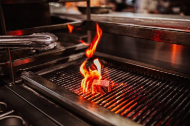 Izgaranın üzerinde canlı alevlerin yükseldiği bir bifteğin detaylı yakın çekim görüntüsü cezbedici bir atmosfer yaratır.