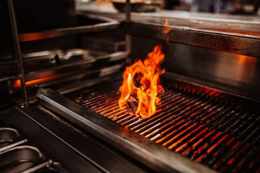 Izgaranın üzerinde canlı alevlerin yükseldiği bir bifteğin detaylı yakın çekim görüntüsü cezbedici bir atmosfer yaratır.