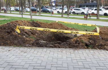 Toprağa kazılmış kare bir çukur, kamu kaldırımının yanında ve tehlike işareti olarak sarı bir kurdele ile çevrili. Kamu hizmeti işleri, yeraltı tesislerinin tamiri.