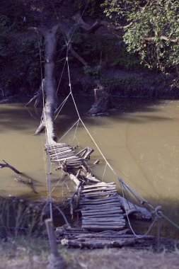 Halatlardan yapılmış korkuluklarla bir nehrin üzerine inşa edilmiş ev yapımı ahşap bir köprü. Tahtalar suya gömülmüş ağaç gövdesine çivilenmiş.