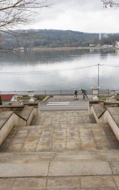 İki insan heykeliyle gölün kenarına inen geniş taş merdiven sakin bir göle ve bulutlu havada orman yamacına bakıyor.