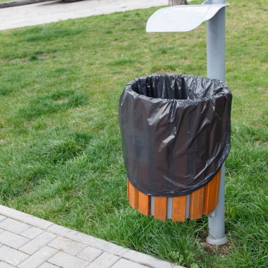 Siyah plastik kaplamalı umumi çöp tenekesi, ahşap kiremitler, kaldırımın yanındaki çimenli park arka planında. Kentsel temizlik kavramı