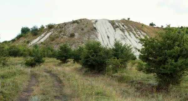 Doğal tebeşir tepesi, görünür erozyon desenleriyle, yabani çalılar ve çimlerle çevrili, kırsal alanda bulutlu bir gökyüzünün altında