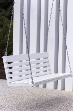 Beyaz ahşap bir salıncak zincire asılı dikey beyaz panellere karşı. Zemin küçük çakıl taşlarıyla kaplı, sakin bir açık hava ortamı yaratıyor.
