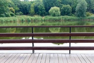 Bir ahşap korkuluk yemyeşil ağaçlarla çevrili sakin bir göle bakar. Su yeşilliği yansıtır, sakin doğal bir manzara yaratır.