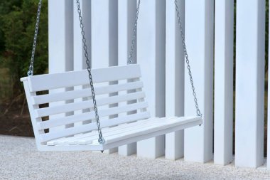 Beyaz ahşap bir salıncak zincire asılı dikey beyaz panellere karşı. Zemin küçük çakıl taşlarıyla kaplı, sakin bir açık hava ortamı yaratıyor.
