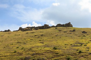 Kayalık bir yamaç, beyaz bulutlu mavi gökyüzünün zeminine doğru yükselir. Çimenler gür ve yeşildir. Gün ortasında doğal ortamda canlı bir kontrastlık yaratır. Güzel manzara.
