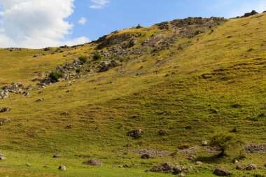 Toprakta yemyeşil tepeler uzanır, kayalık oluşumlarla benekli. Pofuduk bulutlu mavi bir gökyüzü huzurlu bir arka plan sağlar, huzurlu bir kırsal atmosfer yaratır.