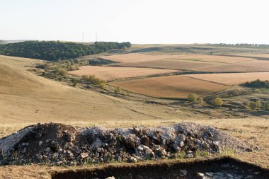 Yuvarlanan tepeler, dağınık kayalar ve çimen parçalarından oluşan sakin bir manzara. Ağaçlar, temiz bir gökyüzünün altında, huzurlu bir doğal ortamı işaret ediyor..