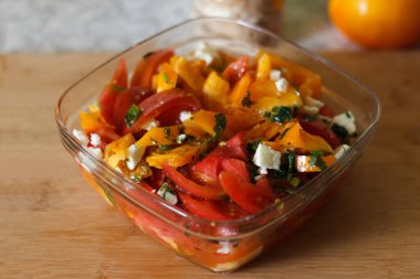 Dilimlenmiş domatesli taze bir salata, sarı dolma biber ve cam kasede parçalanmış peynir. Salata otlarla süslenir ve ahşap bir yüzeyde servis edilir.