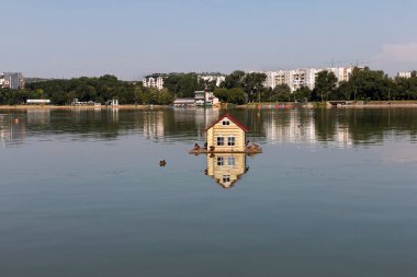Kuşlar için küçük bir ev - kırmızı çatılı bir besleyici kısmen sakin sulara gömülür. Vahşi ördekler dinlenir ve evde yemek yerler. Bir şehrin arka planına karşı