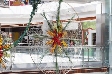 Renkli çiçekler ve yeşilliklerle süslenmiş, modern bir alışveriş merkezinde sergilenen doğal malzemelerden oluşan büyük bir dekoratif çark.