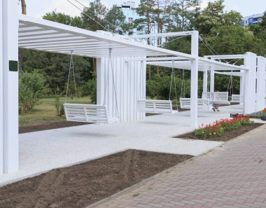 Minimalist bir yapının altında beyaz salıncakların olduğu modern bir açık hava alanı. Mutlu çocukların bindiği. Bölge yeşillik ve çiçek yataklarıyla çevrili.