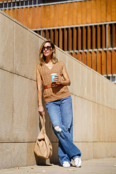 全身上下的成年女子 背着时髦的手提包和外卖的咖啡 靠着现代的墙壁 在阳光灿烂的城市街道上遥望远方 — 图库照片