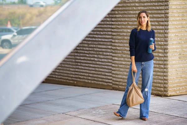全身女性 穿着时髦的牛仔裤和毛衣 头戴手提包和保温瓶 站在现代建筑外的人行道上朝外看 — 图库照片