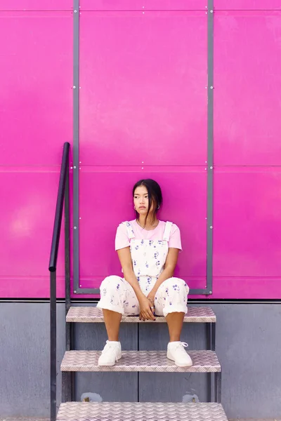 全身上下静谧的年轻亚洲女子 穿着时髦的衣服 靠着粉红的墙壁坐在金属台阶上 望着远方 — 图库照片