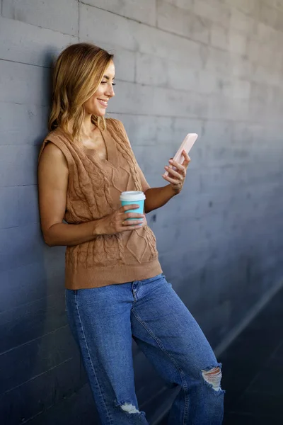 Happy adult female with coffee to go smiling and watching video on cellphone while leaning on gray wall on city street