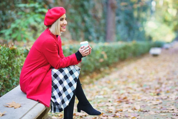 stock image Side view of positive female wearing red beret and coat sitting with legs crossed and having hot takeaway coffee on bench in autumn park