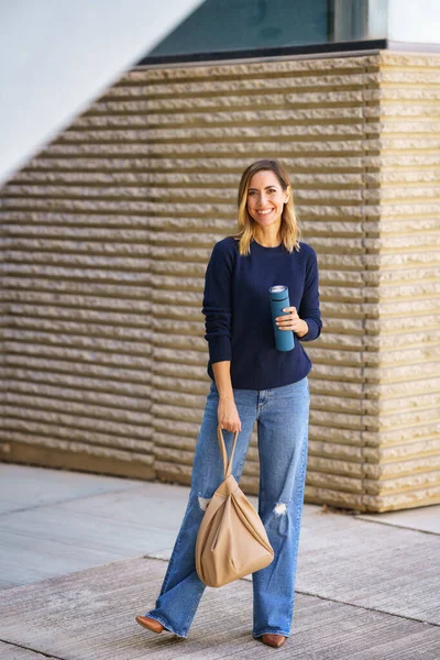 Full body happy female in stylish clothes with thermos and bag smiling and looking at camera while standing on city street
