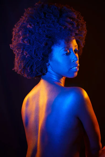 stock image Sensual nude African American female with curly hair looking away over shoulder while standing under colorful neon illumination against black background