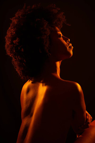 Naked African American woman model with Afro hairstyle closing eyes and enjoying bright neon orange light against black background