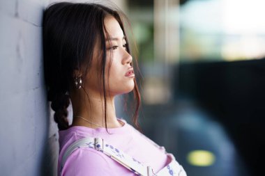 Genç Asyalı kadın örgü örmüş, duvara yaslanmış ve gündüz vakti şehir sokağına bakıyor.