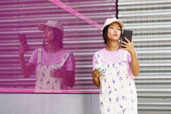 都会のピンクのガラスの壁の近くに立っている間 携帯電話でコーヒー読書メッセージのテイクアウトカップとスタイリッシュな衣装の若いアジアの女性 — ストック写真
