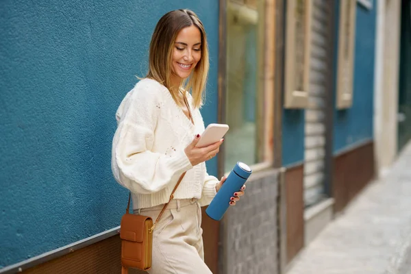 携帯電話やメッセージングを閲覧しながら スマートカジュアルウェアで陽気な若い女性が青い壁の近くに立って — ストック写真