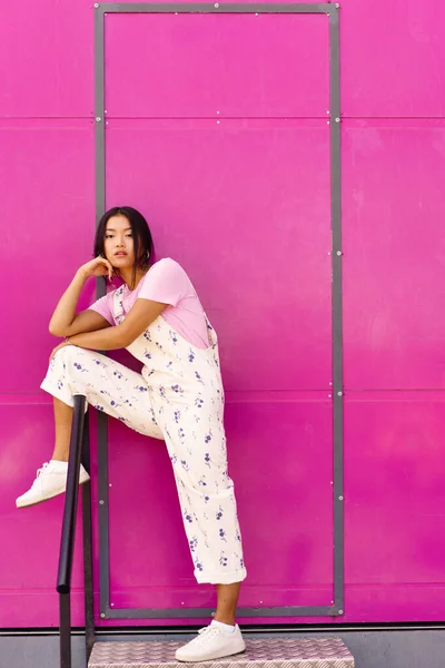 全身年轻的亚洲女子 穿着时髦的衣服 脚踏在栏杆上 手扶着手 脚踏着粉红的墙壁 站在金属台阶上 — 图库照片