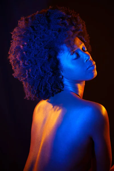 Topless black woman with Afro hairstyle closing eyes under orange and blue neon light against black background