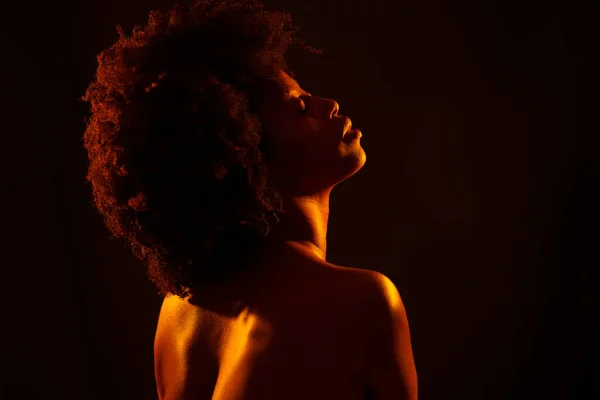 Naked black woman model with Afro hairstyle closing eyes and enjoying bright neon orange light against black background